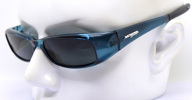 May include: Teal-colored sunglasses with dark lenses. The frames have a glossy finish and a small silver emblem on the temples. The arms are black. The sunglasses are set against a white background.