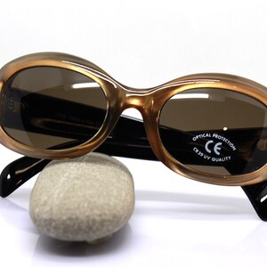 May include: Brown oval-shaped sunglasses with black arms and brown lenses. The sunglasses have "OPTICAL PROTECTION CE CR39 UV QUALITY" printed on the right lens.