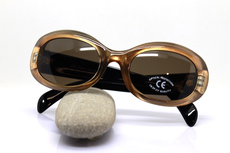 May include: Brown oval-shaped sunglasses with black arms and brown lenses. The sunglasses have "OPTICAL PROTECTION CE CR39 UV QUALITY" printed on the right lens.