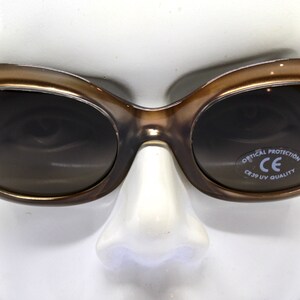 May include: Brown oval-shaped sunglasses with dark lenses. The sunglasses have a CE mark and the text "Optical Protection CE 3.1 UV Quality" on the right lens.