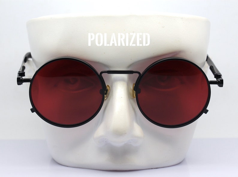 K&ouml;nnte beinhalten: Runde Sonnenbrille mit roten Gl&auml;sern und schwarzem Gestell. Das Wort "POLARIZED" ist wei&szlig; &uuml;ber der Brille aufgedruckt. Die Sonnenbrille hat ein einzigartiges Design mit Steg und Scharnieren.