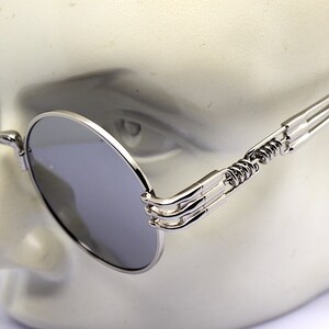 K&ouml;nnte beinhalten: Silberne runde Sonnenbrille mit einem einzigartigen Feder-Scharnier-Design. Die Sonnenbrille hat eine dunkelgraue Linse und einen silbernen Metallrahmen.