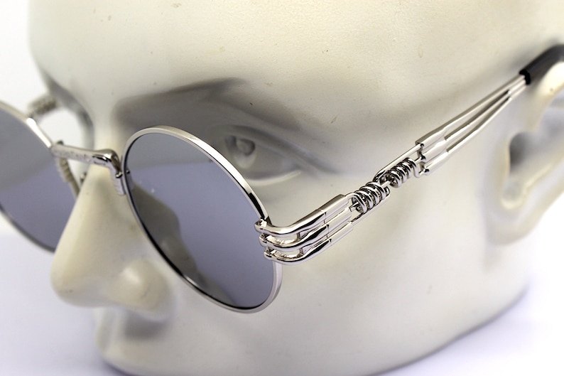 K&ouml;nnte beinhalten: Silberne runde Sonnenbrille mit einem einzigartigen Feder-Scharnier-Design. Die Sonnenbrille hat eine dunkelgraue Linse und einen silbernen Metallrahmen.