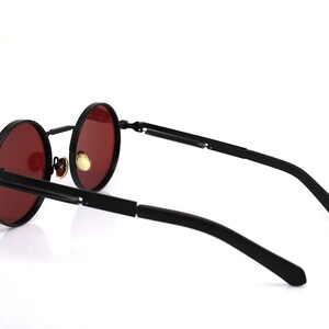 K&ouml;nnte beinhalten: Schwarze runde Sonnenbrille mit roten Gl&auml;sern. Die Sonnenbrille hat einen schwarzen Rahmen und B&uuml;gel. Die Gl&auml;ser sind tiefrot. Die B&uuml;gel haben ein einzigartiges Design mit einem Scharnier.