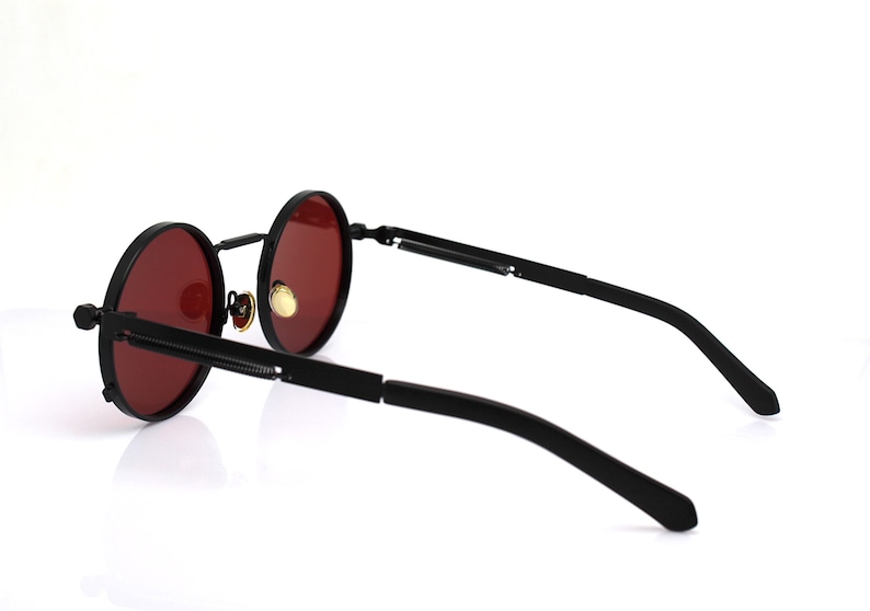 K&ouml;nnte beinhalten: Schwarze runde Sonnenbrille mit roten Gl&auml;sern. Die Sonnenbrille hat einen schwarzen Rahmen und B&uuml;gel. Die Gl&auml;ser sind tiefrot. Die B&uuml;gel haben ein einzigartiges Design mit einem Scharnier.