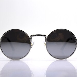 K&ouml;nnte beinhalten: Silberne runde Sonnenbrille mit dunkelgrauen Gl&auml;sern. Die Sonnenbrille hat einen detaillierten Metallrahmen.