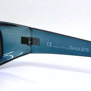 May include: Close-up of teal sunglasses with dark lenses. The frame is a glossy teal color. The text "CE MADE IN ITALY by DANOR 675" is printed on the arm of the sunglasses. The sunglasses are likely for eye protection.