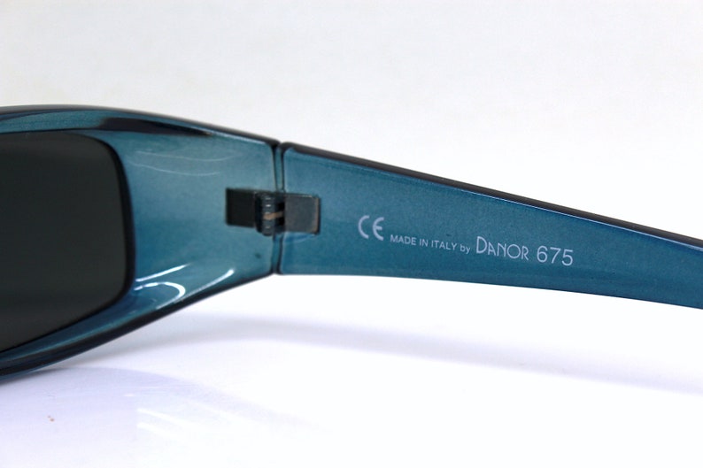 May include: Close-up of teal sunglasses with dark lenses. The frame is a glossy teal color. The text "CE MADE IN ITALY by DANOR 675" is printed on the arm of the sunglasses. The sunglasses are likely for eye protection.