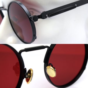 K&ouml;nnte beinhalten: Nahaufnahme einer runden Sonnenbrille mit schwarzen Rahmen und roten Gl&auml;sern. Die Rahmen haben ein Federscharnier-Design und goldfarbene Akzente. Die Gl&auml;ser sind tiefrot.