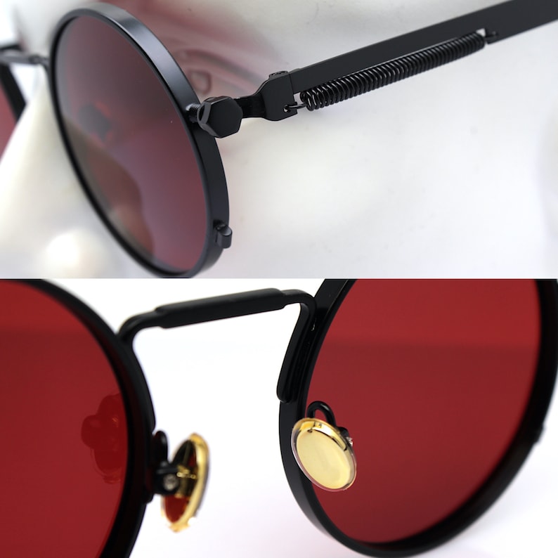 K&ouml;nnte beinhalten: Nahaufnahme einer runden Sonnenbrille mit schwarzen Rahmen und roten Gl&auml;sern. Die Rahmen haben ein Federscharnier-Design und goldfarbene Akzente. Die Gl&auml;ser sind tiefrot.