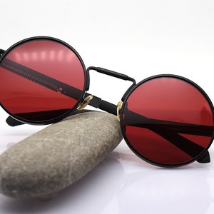 K&ouml;nnte beinhalten: Runde, schwarze Sonnenbrille mit roten Gl&auml;sern. Die Brille hat ein ausgepr&auml;gtes Steampunk-Design mit Steg und Scharnieren. Die Sonnenbrille liegt auf einem grauen Stein vor wei&szlig;em Hintergrund.
