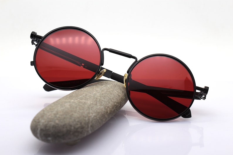 K&ouml;nnte beinhalten: Runde, schwarze Sonnenbrille mit roten Gl&auml;sern. Die Brille hat ein ausgepr&auml;gtes Steampunk-Design mit Steg und Scharnieren. Die Sonnenbrille liegt auf einem grauen Stein vor wei&szlig;em Hintergrund.