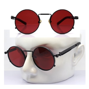 K&ouml;nnte beinhalten: Runde Sonnenbrille mit schwarzem Gestell und roten Gl&auml;sern. Die Brille hat ein klassisches, Vintage-Design. Die Gl&auml;ser sind tiefrot und das Gestell ist mattschwarz. Die Sonnenbrille wird auf einem wei&szlig;en Schaufensterkopf gezeigt.