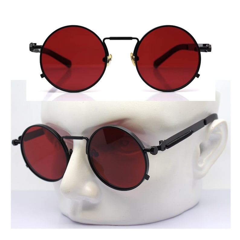 K&ouml;nnte beinhalten: Runde Sonnenbrille mit schwarzem Gestell und roten Gl&auml;sern. Die Brille hat ein klassisches, Vintage-Design. Die Gl&auml;ser sind tiefrot und das Gestell ist mattschwarz. Die Sonnenbrille wird auf einem wei&szlig;en Schaufensterkopf gezeigt.