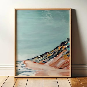 May include: A framed abstract painting of a beach scene. The artwork features a light blue sky, a sandy beach, and a colorful hillside. The frame is made of light-colored wood.