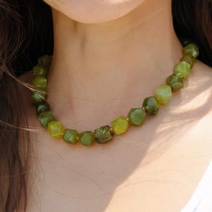 May include: A green beaded necklace with varying shades of green stones. The necklace is made of faceted, irregular-shaped beads strung together with gold-colored string. The necklace is worn against a white tank top.