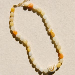 May include: A beaded necklace featuring smooth, rounded stones in shades of cream, pale yellow, and orange. The necklace includes a single white spiral seashell charm and a gold-toned metal chain extender with a lobster clasp closure.