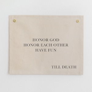 May include: A rectangular beige fabric wall hanging with gold-colored grommets in each corner. The black text reads "HONOR GOD HONOR EACH OTHER HAVE FUN" and "TILL DEATH". The wall hanging is displayed against a white wall.