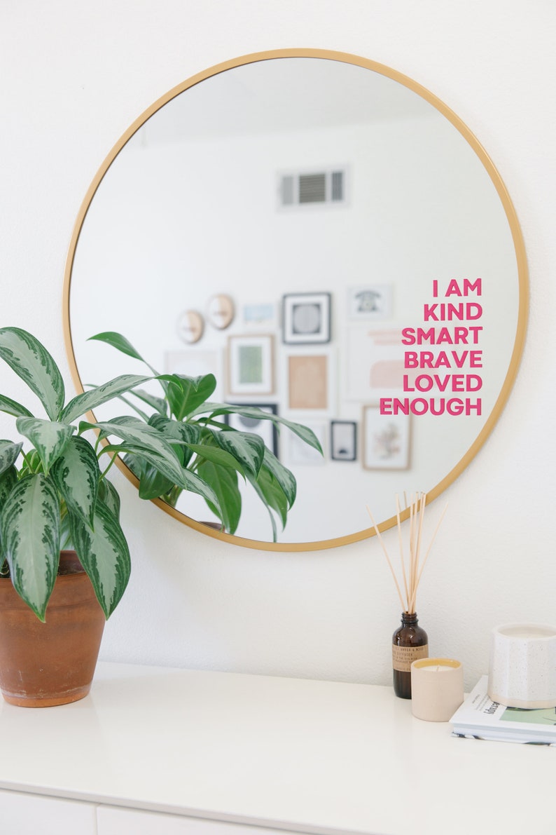 May include: A gold round mirror with a white wall behind it. The mirror reflects a wall with framed artwork and the words "I AM KIND SMART BRAVE LOVED ENOUGH" written in pink on the mirror.