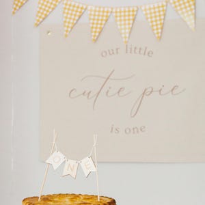 May include: A golden-brown pie in a silver tin, topped with a small banner that reads "ONE". A sign in the background says "our little cutie pie is one". Yellow and white checkered pennant banner.