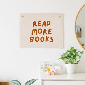May include: A white canvas banner with the text "READ MORE BOOKS" in orange lettering. The banner is hanging on a white wall with a small wooden toy car and a book on a white dresser below.