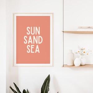 Może przedstawiać: Wydruk w ramie z brzoskwiniowym tłem i tekstem "SUN SAND SEA" w kolorze białym.