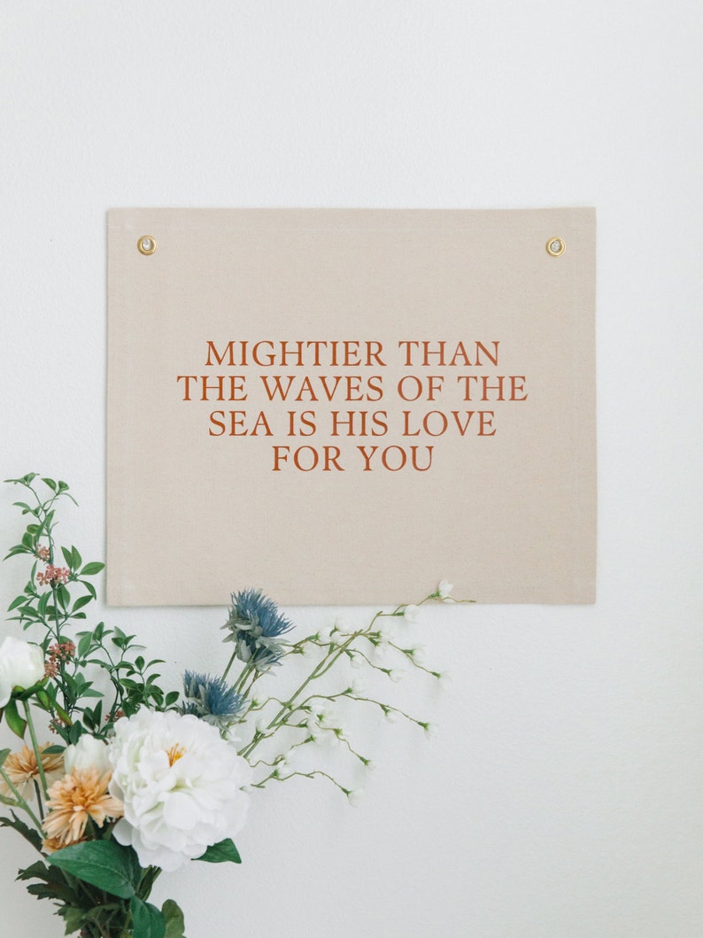 May include: A beige canvas wall tapestry with the text "MIGHTER THAN THE WAVES OF THE SEA IS HIS LOVE FOR YOU" printed in brown.