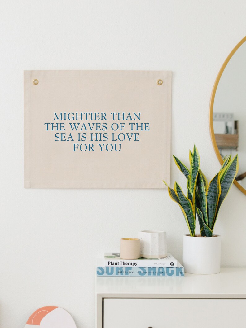 May include: A beige canvas wall tapestry with the text "MIGHTER THAN THE WAVES OF THE SEA IS HIS LOVE FOR YOU" in blue lettering. The tapestry is hanging on a white wall above a white dresser with a plant, a book, and two decorative containers.