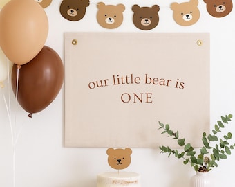 May include: A party scene with a banner that reads "our little bear is ONE." The banner is decorated with bear-shaped cutouts. There are balloons, a cake with a bear topper, and snacks. The color scheme is neutral with brown and beige tones.