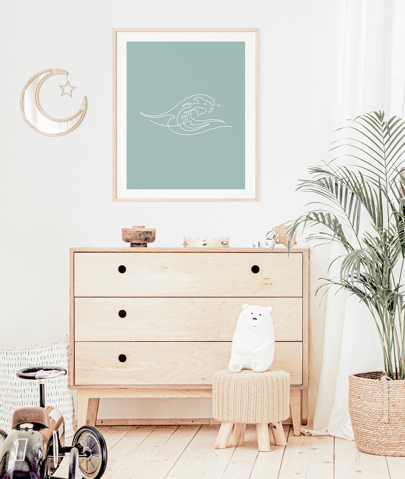 May include: A framed print with a minimalist white wave design on a teal background. The artwork is displayed above a wooden chest of drawers. A crescent moon mirror and a white bear figurine are also visible.