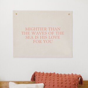 May include: A beige canvas wall tapestry with the text "Mightier than the waves of the sea is his love for you" printed in pink.