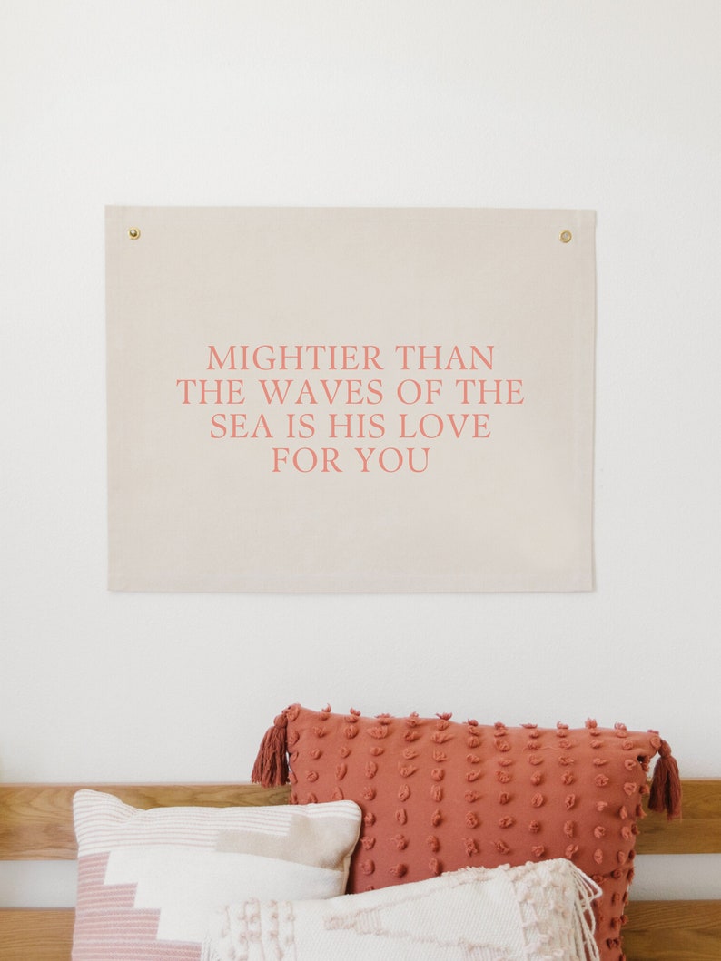 May include: A beige canvas wall tapestry with the text "Mightier than the waves of the sea is his love for you" printed in pink.