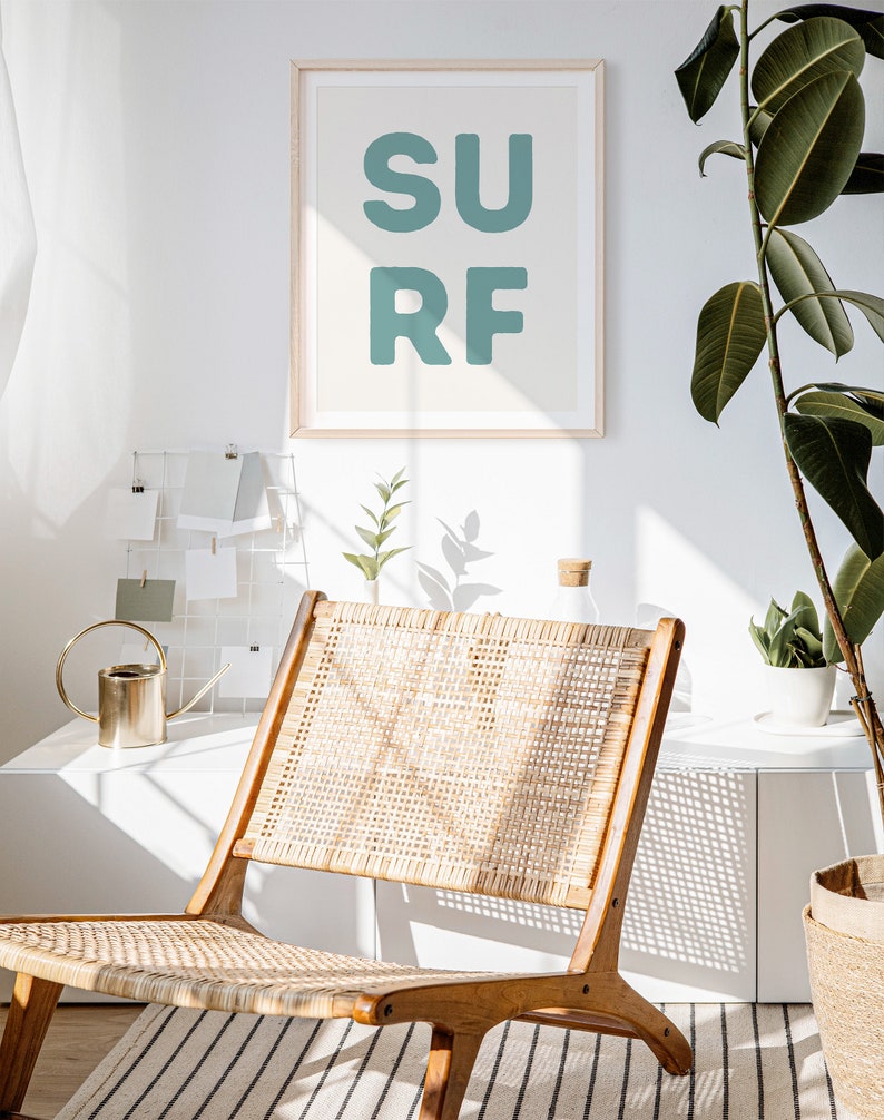 May include: A framed print with the word "SURF" in teal letters hangs above a wooden chair with a woven seat. The room is bright with a white wall and a large potted plant. A small gold watering can sits on a white surface.