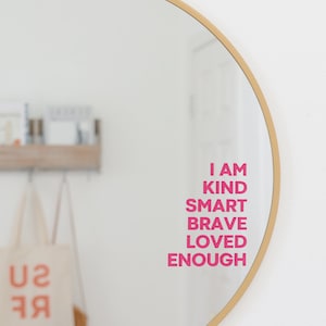 May include: A round gold-framed mirror with the words "I am kind smart brave loved enough" written in pink on the reflection.