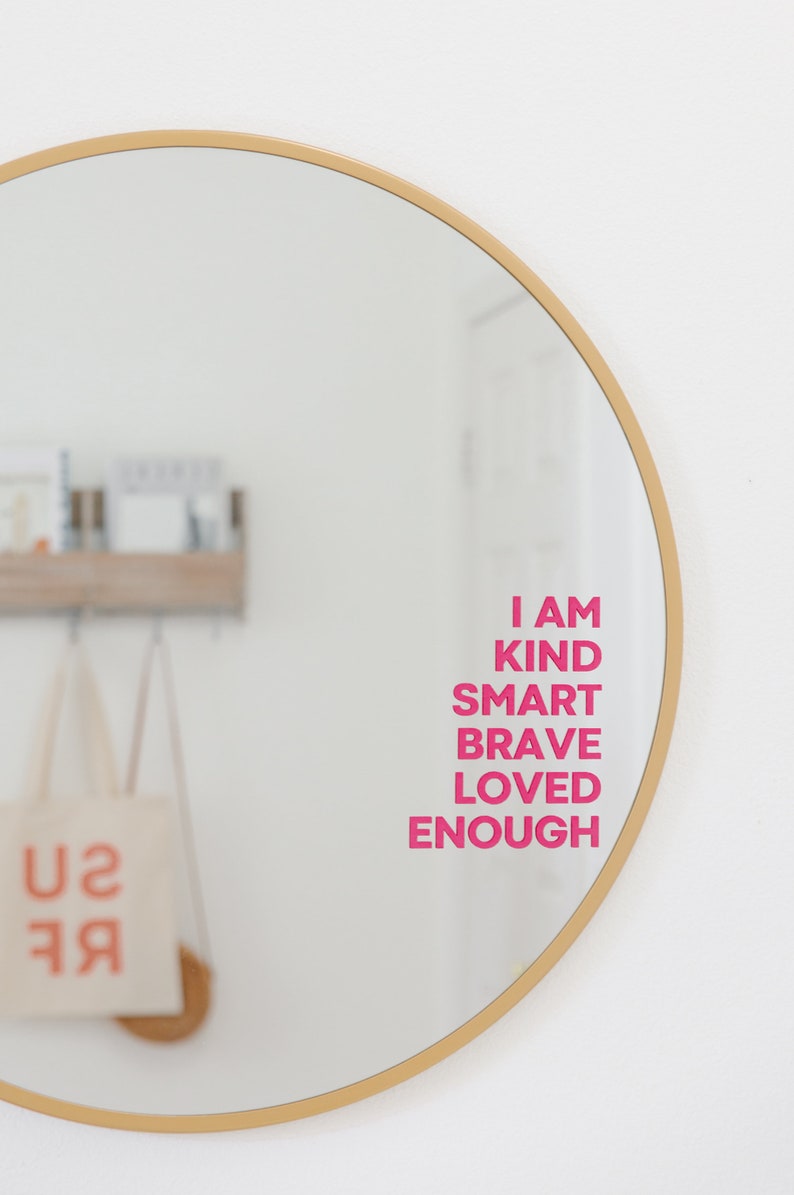 May include: A round gold-framed mirror with the words "I am kind smart brave loved enough" written in pink on the reflection.