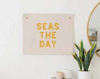 May include: A rectangular beige canvas wall hanging featuring the mustard yellow text SEAS THE DAY in a bold sans-serif font. The fabric banner includes two metallic gold grommets at the top corners for mounting purposes on a flat surface.