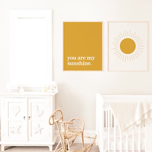 May include: Two framed art prints with a nursery theme. One is mustard yellow with the text "you are my sunshine." The other features a yellow sunburst design on a cream background. The prints are displayed above a white cot.
