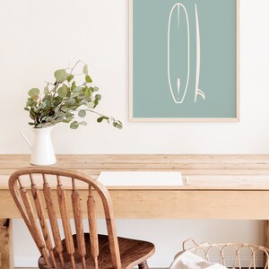 May include: A framed print with a minimalist surfboard design in white on a teal background. The print is on a wooden desk with a wooden chair, a white jug with greenery, and a woven basket. The scene has a coastal, relaxed feel, with a light wood desk.