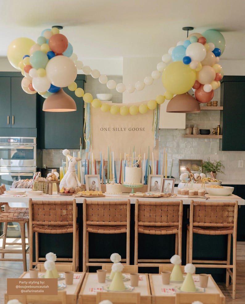 May include: A birthday party table setting with a white cake, a banner that reads "One Silly Goose", and a balloon arch in pastel colors. The table is decorated with candles, a stuffed bunny, and framed photos.