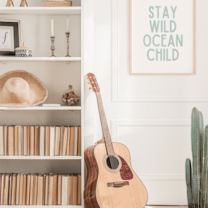 May include: Acoustic guitar leaning against a white wall next to a bookshelf and a framed print that reads "Stay Wild Ocean Child". A cactus in a terracotta pot is on the right. The room has a neutral colour palette.