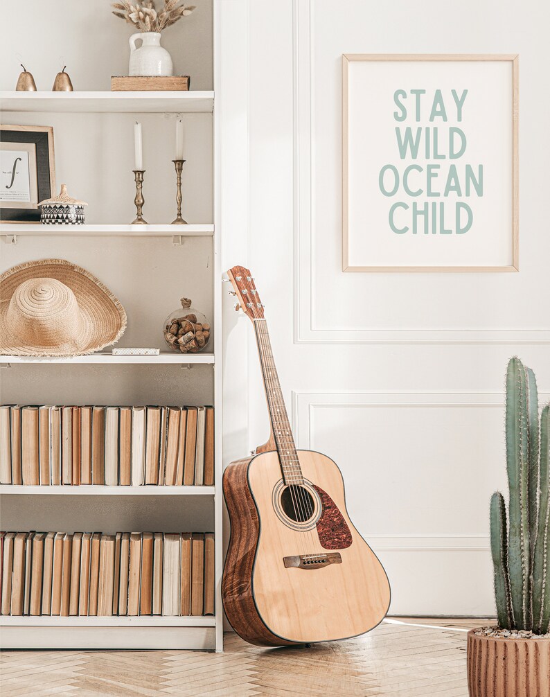 May include: Acoustic guitar leaning against a white wall next to a bookshelf and a framed print that reads "Stay Wild Ocean Child". A cactus in a terracotta pot is on the right. The room has a neutral colour palette.