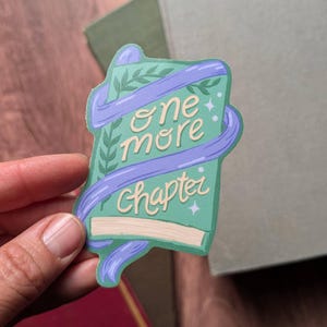 May include: A sticker with a green book design, wrapped with a purple ribbon. The text "one more chapter" is written on the book in a whimsical font. The sticker has a light green background with small stars and leaf accents.