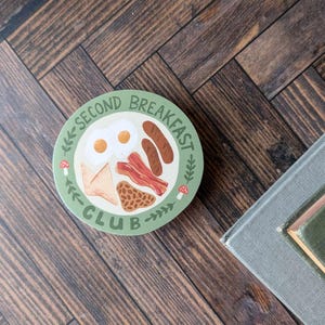 May include: A green round sticker with the text "Second Breakfast Club" and a cartoon illustration of a plate of breakfast food, including eggs, bacon, sausage, and toast.