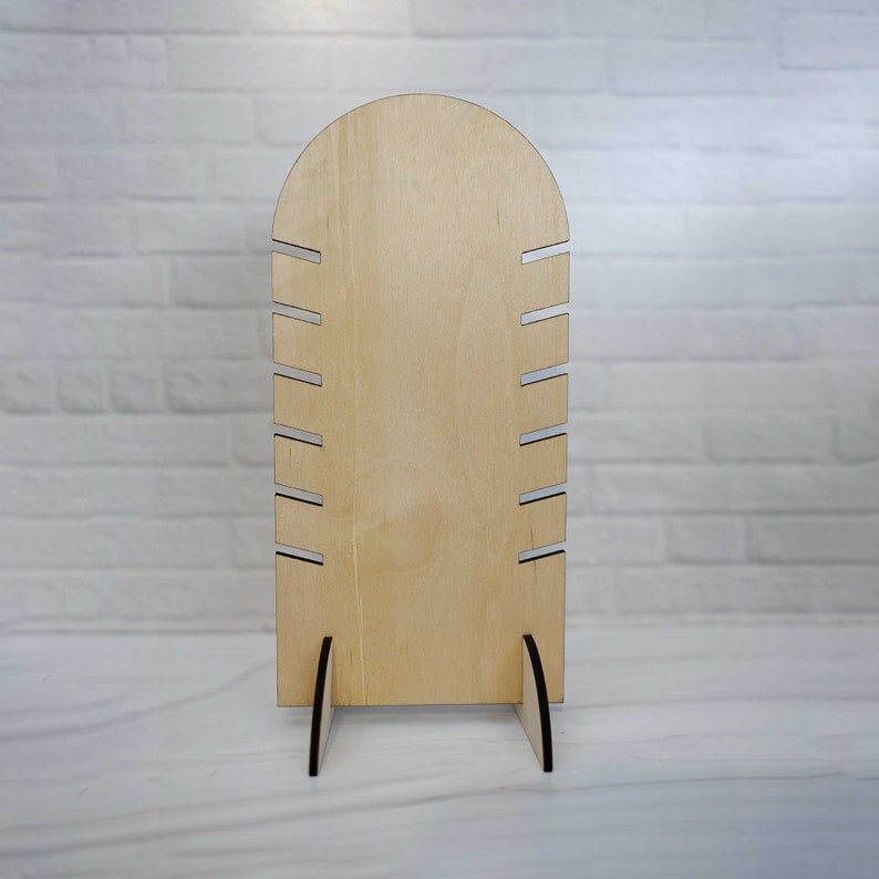 May include: A wooden jewelry display stand with an arched top and multiple slots for holding necklaces or bracelets. The stand is made of natural wood and has a light brown color.