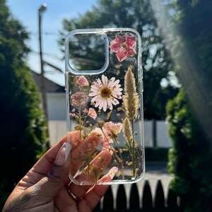 May include: A clear phone case with pressed flowers and gold flakes. The flowers are pink, white, and brown. The case is designed to fit a smartphone.