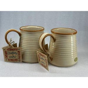 May include: Two white ceramic mugs with brown rims and a brown stripe pattern. The mugs are sitting on a table with a brown and white tag that says "Cranbury Crafts".