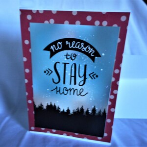 May include: A greeting card with a red polka dot border. The card features a blue and white background with the text "no reason to STAY home" in black script. A silhouette of trees is at the bottom.