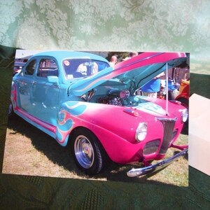 May include: A teal and pink 1940s vintage car with the hood open. The car has flames painted on the side and a chrome bumper.