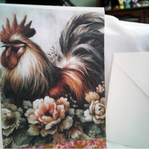 May include: Greeting card featuring a detailed illustration of a rooster with brown, white, and black feathers, surrounded by peach and cream-colored flowers. A white envelope is next to the card. The card has a floral patterned border.
