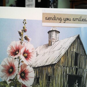 May include: Greeting card featuring a watercolor painting of a weathered barn and hollyhock flowers. The flowers are white with pink and red centers. The text "sending you smiles" is printed on a beige banner.